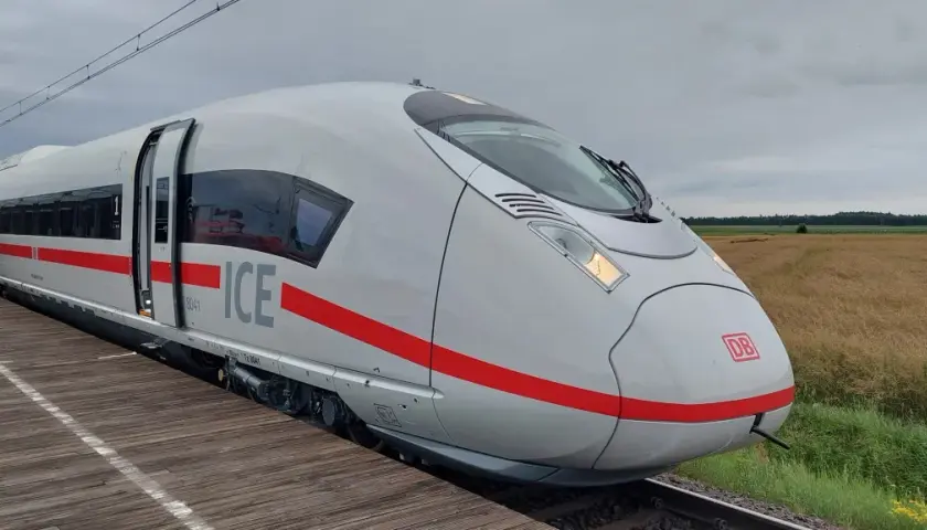 Biały pociąg Siemens Velaro ICE z czerwonym pasem stoi na drewnianym peronie. W tle pole i zachmurzone niebo.