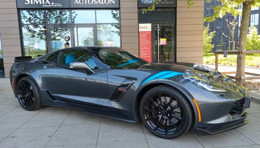 Szary Chevrolet Corvette C7 z niebieskimi pasami, zaparkowany przed salonem samochodowym. Sprawdź aktualną corvette c7 cena!