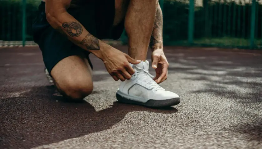 Mężczyzna z tatuażami na ramieniu wiąże białe buty sportowe. Po treningu może pojawić się ból kolan, jak leczyć go skutecznie?