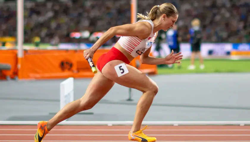 Zawodniczka biegnie z pałeczką w sztafecie 4x100 kobiet. Numer 5 na udzie.
