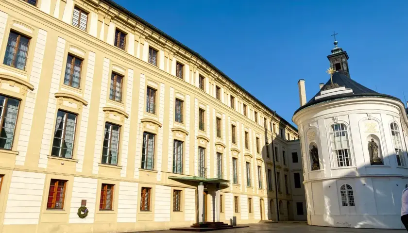 Hradczany zamek w Pradze. Długi, żółty budynek z wieloma oknami i biała, okrągła kaplica pod błękitnym niebem.