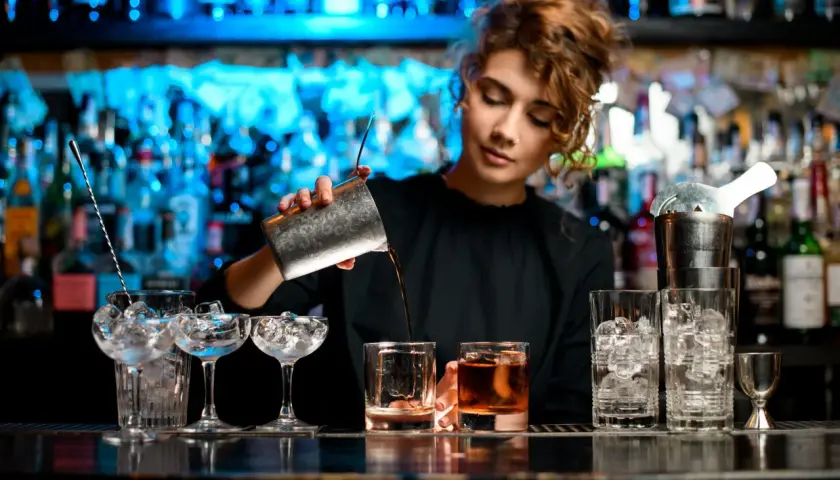Kto robi drinki? Młoda barmanka z kręconymi włosami przelewa płyn z shakera do szklanki z lodem.