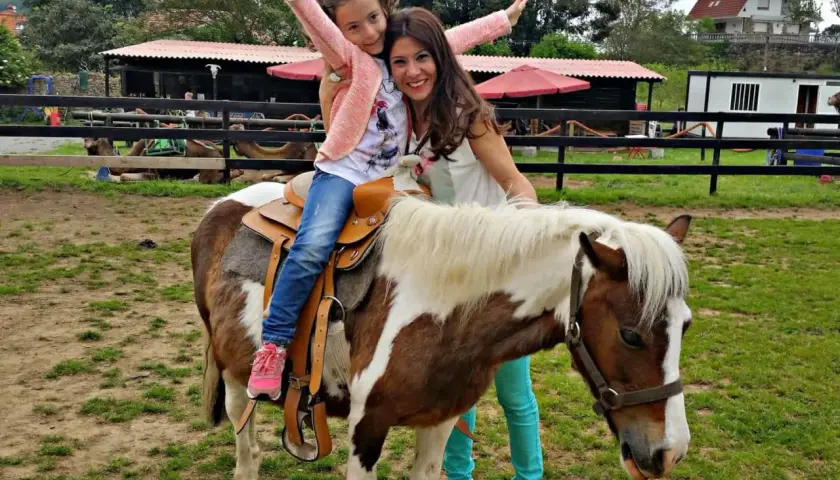 Niña feliz en un poni, disfrutando de una ruta a caballo en Cantabria. Madre sonriente la acompaña.
