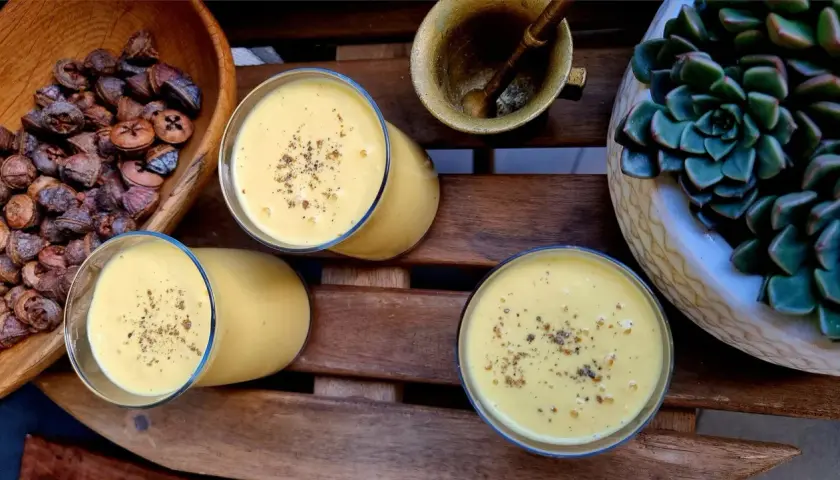 Trzy szklanki kremowego mango lassi, posypane przyprawami, obok miski z suszonymi owocami i sukulentu.