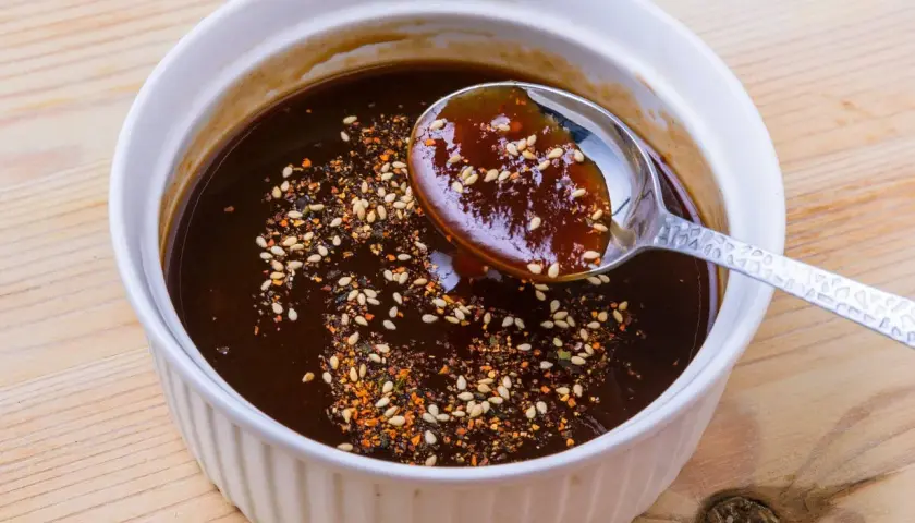 A spoon scoops rich, dark okonomiyaki sauce from a white bowl, sprinkled with sesame seeds and chili flakes. This is how to make okonomiyaki sauce.