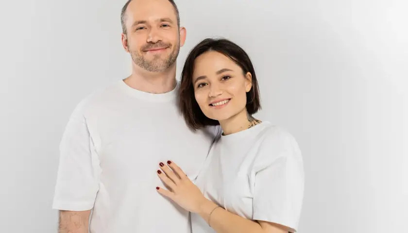 Uśmiechnięta para w białych koszulkach. Ona kładzie dłoń na jego piersi. Pewnie zastanawiają się nad śmiesznymi pytaniami.