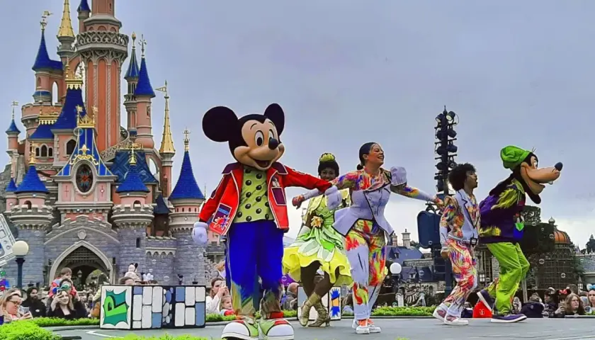 Myszka Miki i Goofy na scenie przed zamkiem w Disneyland Paris, otoczeni tancerzami w kolorowych strojach. Niezapomniane atrakcje Disneyland Paris!