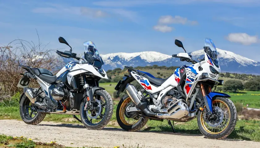 Dos motos de aventura, una Honda Africa Twin 2026 y otra blanca, aparcadas en un camino rural con montañas nevadas al fondo.