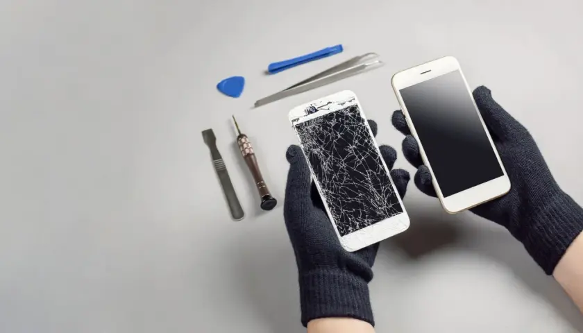 Ręce w rękawiczkach trzymają dwa telefony: jeden z pękniętym ekranem, drugi sprawny. Obok narzędzia do naprawy. Upadek telefonu skutki widoczne.