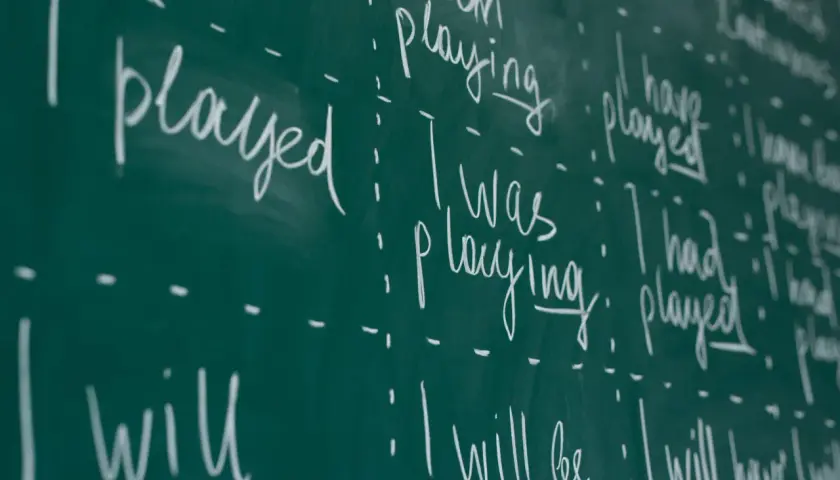 Scrittura a mano su lavagna verde: "I played", "I was playing", "I have played", "I will play". Esercizi di grammatica inglese, con focus sui tempi verbali.