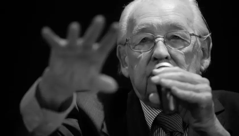 Portret starszego mężczyzny w okularach, trzymającego mikrofon. Wśród znanych reżyserów polskich, jego postać budzi szacunek.