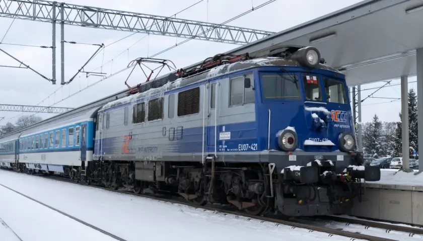 Lokomotywa EU07-421 PKP Intercity z pociągiem na tle zimowego krajobrazu. Widać elementy sieci trakcyjnej i peron.