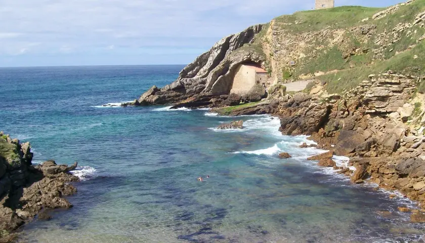 Costa rocosa y mar turquesa en Santillana del Mar, Santander. Una pequeña ermita se esconde en la roca y una torre vigía corona la colina.