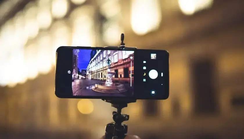 Ręka trzyma smartfon na statywie, jego kamera rejestruje nocną ulicę z fontanną.