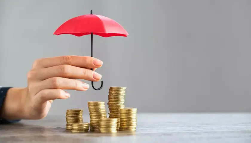 Une main protège des piles de pièces avec un petit parapluie rouge, symbolisant l'assurance PTZ.