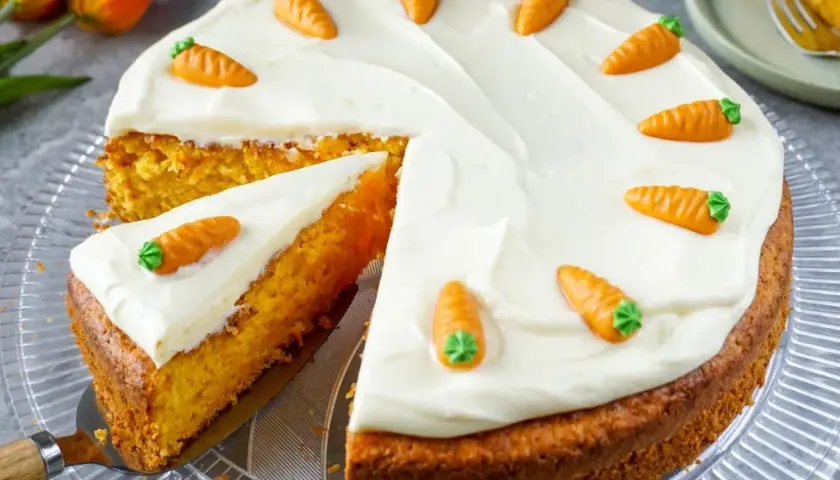 Ein saftiger Karottenkuchen mit cremiger Glasur und süßen Marzipan-Karotten. Ein Stück wurde bereits angeschnitten.