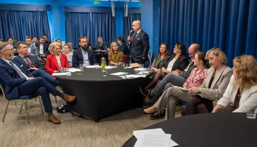 Grupa ludzi siedzi przy okrągłych stołach podczas spotkania, jeden mężczyzna przemawia.