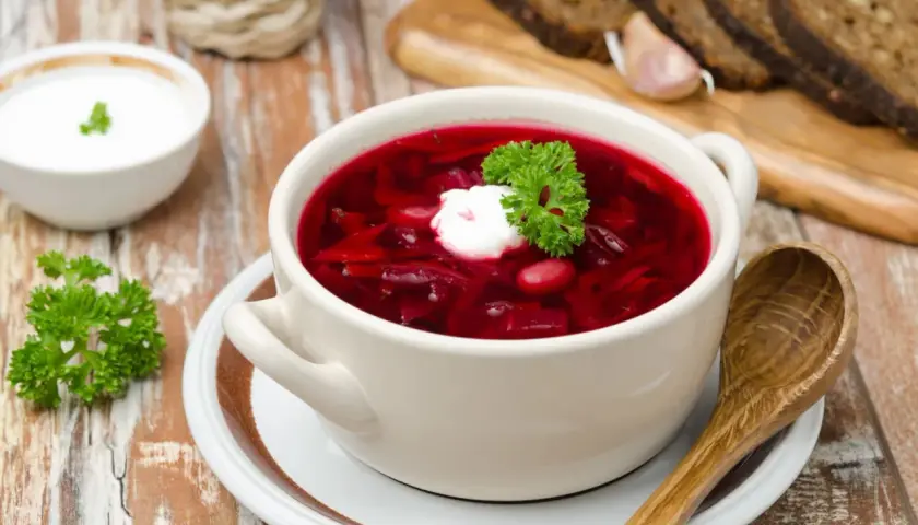 Pyszny botwinka przepis na rozgrzewającą zupę z buraków, podana z kleksem śmietany i natką pietruszki. Obok kromki chleba i czosnek.