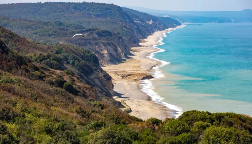 Widok na wybrzeże Karaburun z klifami, plażą i turkusowym morzem. W oddali widać paralotnię.