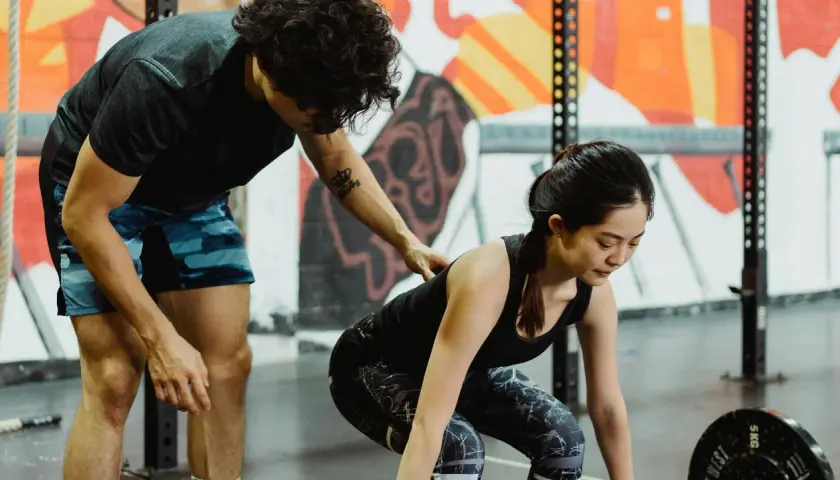 Entrenador ayuda a mujer a levantar pesas en gimnasio con mural colorido de fondo.