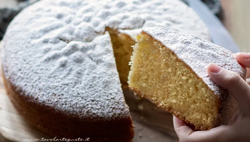 Una fetta di torta margherita ricetta originale, soffice e cosparsa di zucchero a velo, viene sollevata da una mano.