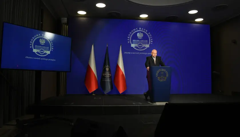 Prezes NBP Adam Glapiński przemawia na tle flag i ekranu z logo banku. Wiron 1m.