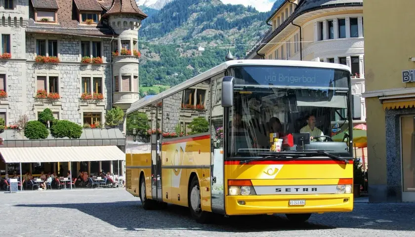Żółty bus do Szwajcarii, z napisem "via Brigerbad", stoi na brukowanym placu przed kamiennymi budynkami i górami.