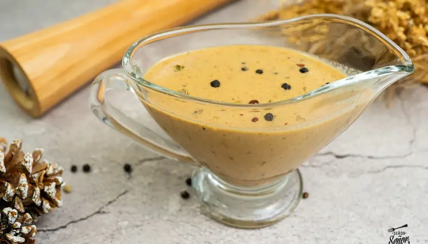 Salsa a la pimienta cremosa en una salsera de cristal, con granos de pimienta negra esparcidos.
