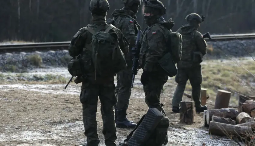 Żołnierze w pełnym rynsztunku, w tym kapitan major, stoją przy torach kolejowych.