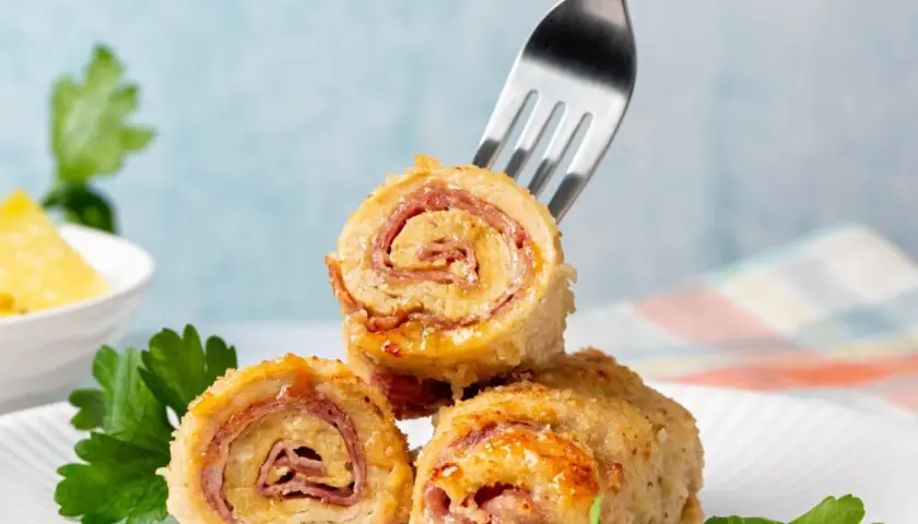 Roladki drobiowe z serem, zawinięte z szynką, prezentują się apetycznie na białym talerzu. Jedna z nich jest nabita na widelec.