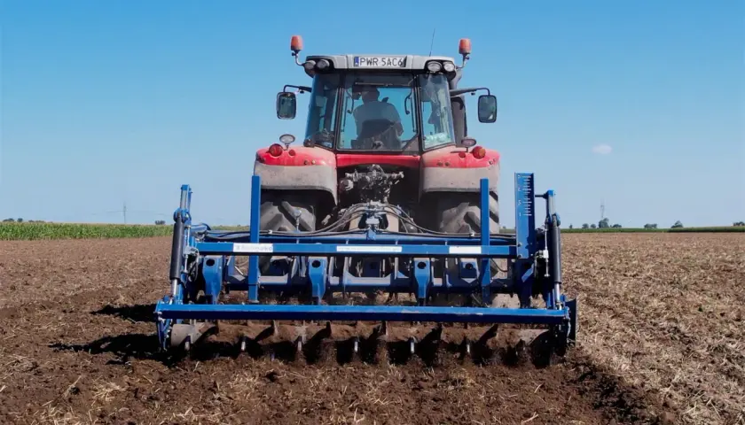 Traktor mit einem blauen Gerät für pfluglose Bodenbearbeitung auf einem Feld unter blauem Himmel.