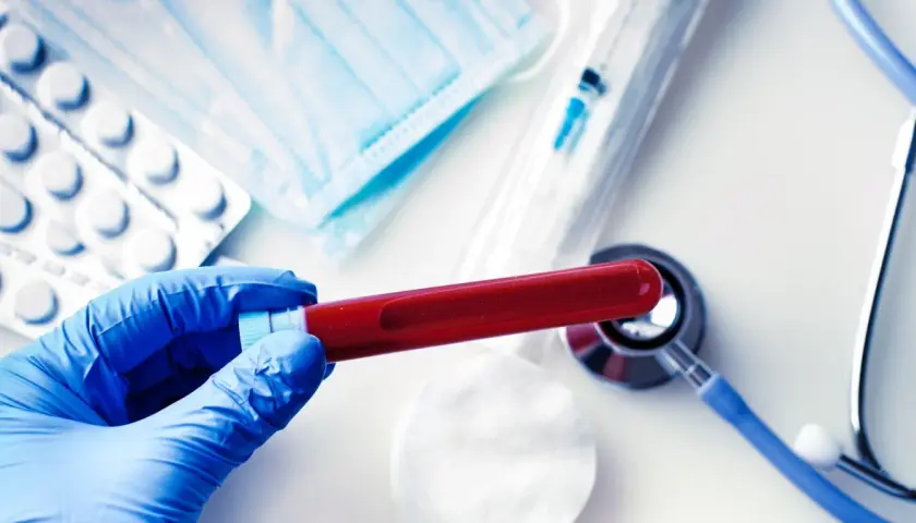 Rękawiczka w niebieskiej rękawicy trzyma probówkę z krwią. Obok leżą maseczka, tabletki, strzykawka i stetoskop. Wyniki badań wskazują, że cholesterol jest podwyższony.