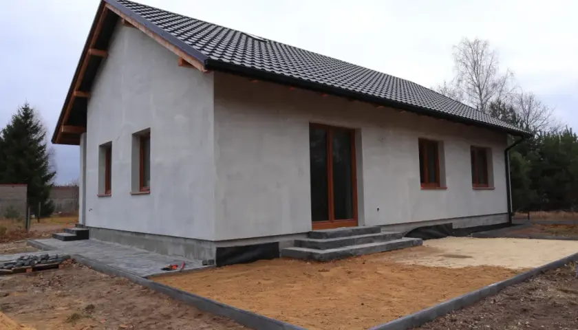 Nowy dom w trakcie budowy, z szarym tynkiem i brązowymi oknami. Wniosek o warunki zabudowy złożony, teren wokół domu przygotowany do dalszych prac.