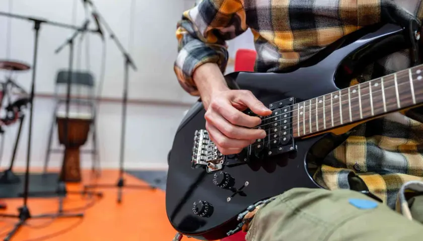 Ręce muzyka grające na czarnej gitarze elektrycznej. W tle stojaki z mikrofonami i bęben. To może być początek nauki, jak zmienić struny w gitarze elektrycznej.