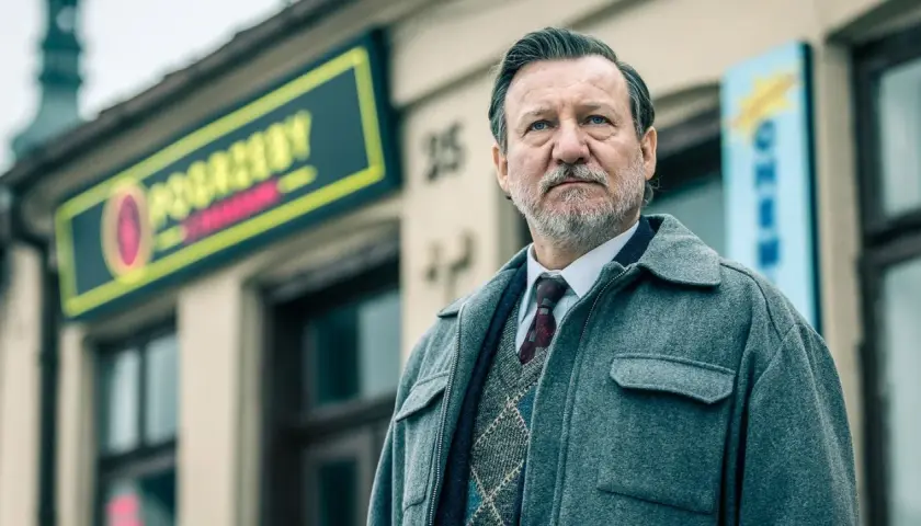 Mężczyzna w eleganckim swetrze i płaszczu, z siwą brodą, stoi przed budynkiem z neonem. To jeden z aktorów z obsady "Minuta ciszy".