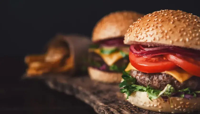 Dwa soczyste smashburgery z serem, pomidorem i czerwoną cebulą, obok kubełka z frytkami.