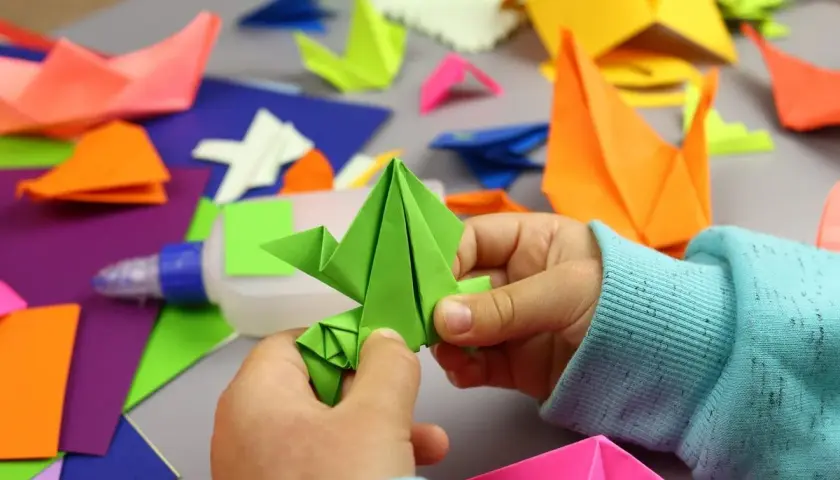 Dziecko tworzy proste origami z kartki a4. Wokół kolorowe figurki i klej.