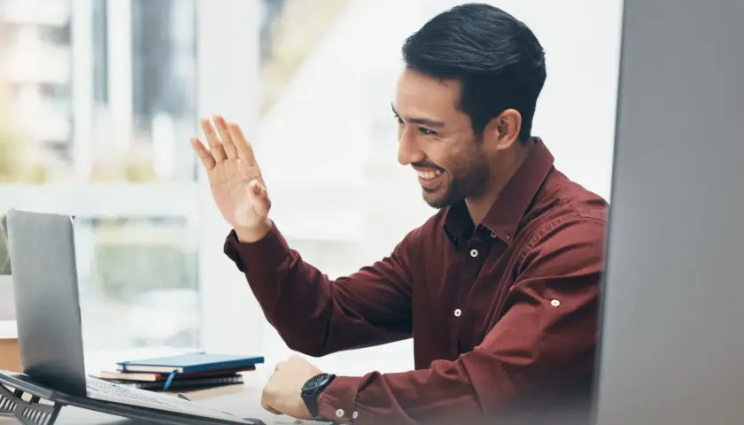 Man waves hello during an online emotional intelligence coaching session, smiling warmly at his laptop.