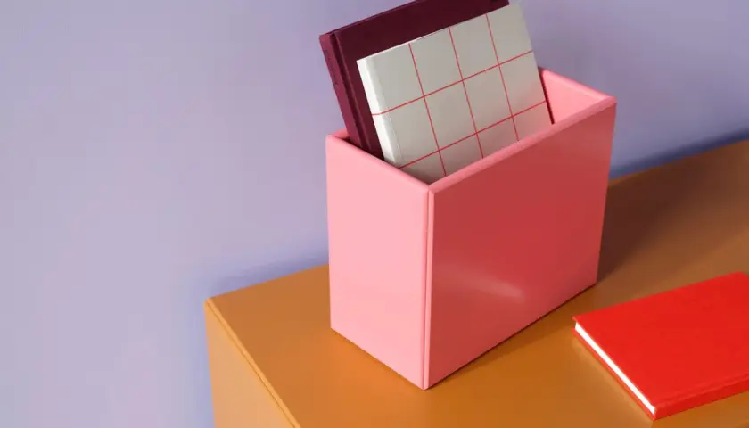 Ein rosa Behälter mit Büchern steht auf einem orangefarbenen Regal vor einer lila Wand. Welche Farbe passt zu lila? Hier sehen wir Rosa und Orange als passende Akzente.