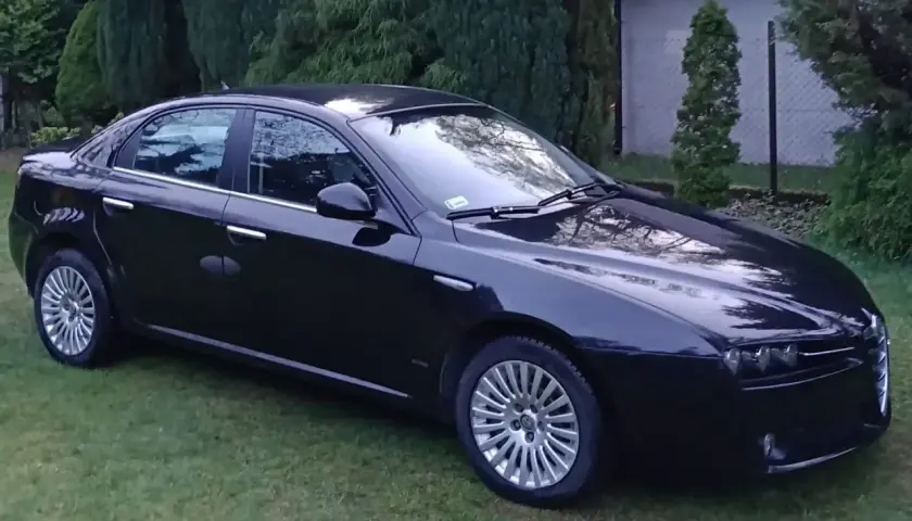 Czarna Alfa Romeo 159 lśni na trawie, otoczona zielenią drzew.