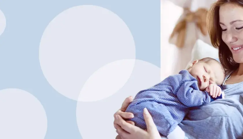 Uśmiechnięta mama tuli śpiącego noworodka w pasiastym pajacyku. Idealny strój na pierwsze wyjście ze szpitala.