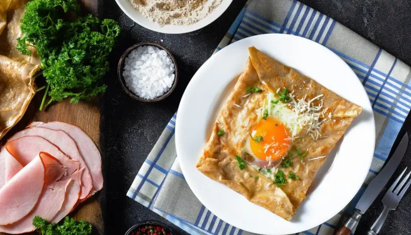 Francuska crepe z jajkiem sadzonym, serem i natką pietruszki na talerzu, obok szynka i składniki.