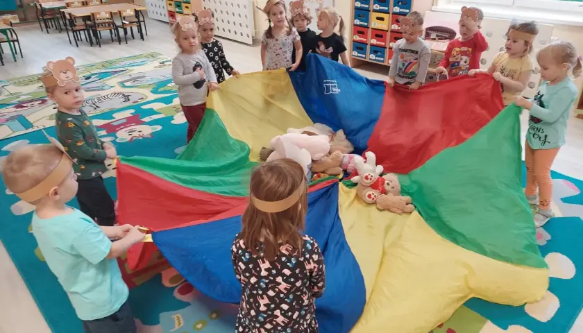 Dzieci w opaskach z uszami misia bawią się kolorowym spadochronem z pluszakami.