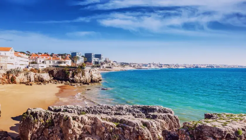 Felsige Küste mit Sandstrand und weißen Häusern, Blick auf das türkisfarbene Meer und die Skyline von Lissabon. Ein perfekter lissabon strand.