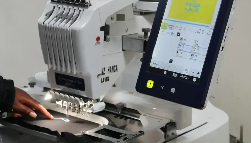 Une machine à broder professionnelle, type impression DTG, crée un logo sur un tissu. L'écran tactile affiche les détails du projet.