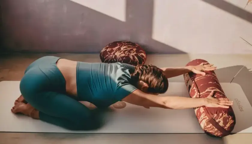 Kobieta w pozycji balasana na macie do jogi, z rękami wyciągniętymi do przodu i podpartymi wałkiem.