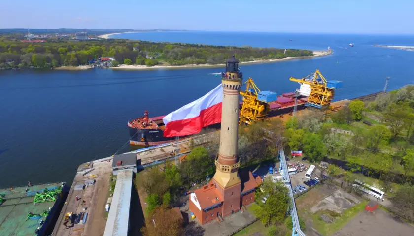 Najwyższa latarnia w Polsce z polską flagą, obok statku i portowych dźwigów.