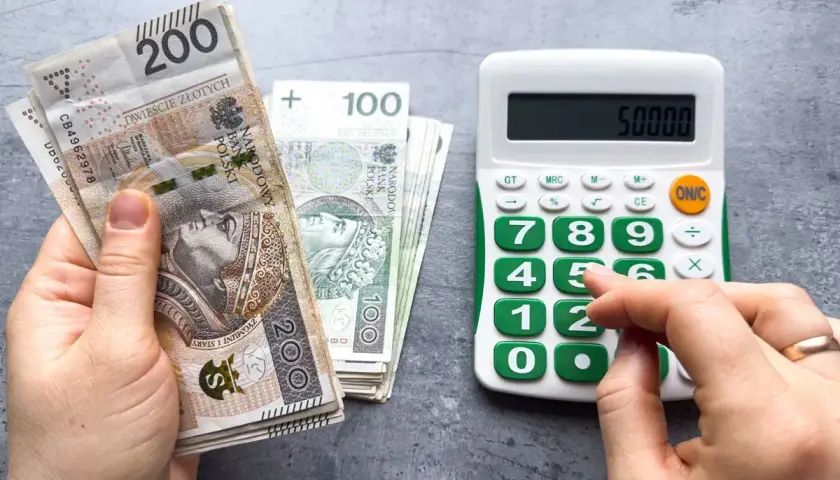 Ręka trzyma polskie banknoty 200 zł i 100 zł, obok kalkulator z wyświetloną kwotą 50000. Obliczenia pokazują, ile pracodawca płaci za pracownika przy najniższej krajowej.