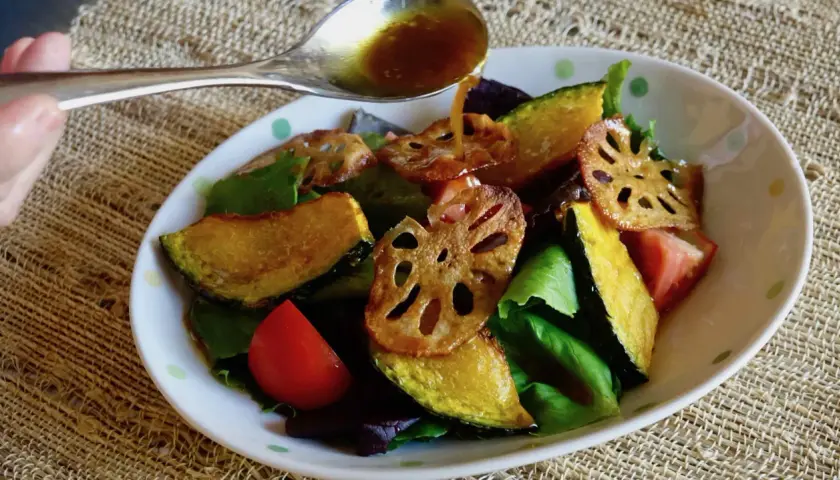 A spoon pours a golden dressing over a salad of roasted pumpkin, lotus root, tomatoes, and greens. The dressing, with a hint of wasabi, adds a zesty kick.