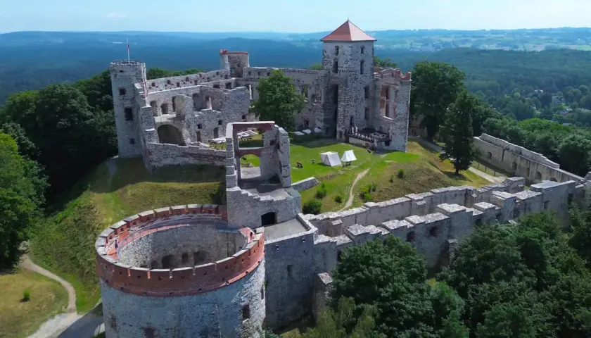 Ruiny zamku Tenczyn na wzgórzu, otoczone zielenią. Widać wieże, mury obronne i dziedziniec z namiotami.